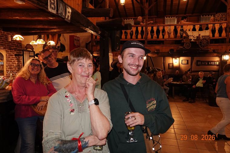 Lachende bezoekers met een glas in de hand in de saloon, onder het bordje BAR